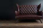 Sofa of black leather standing in center on concrete floor against dark grey wall with copy space. Vintage brown leather sofa with grunge gray wall living room.